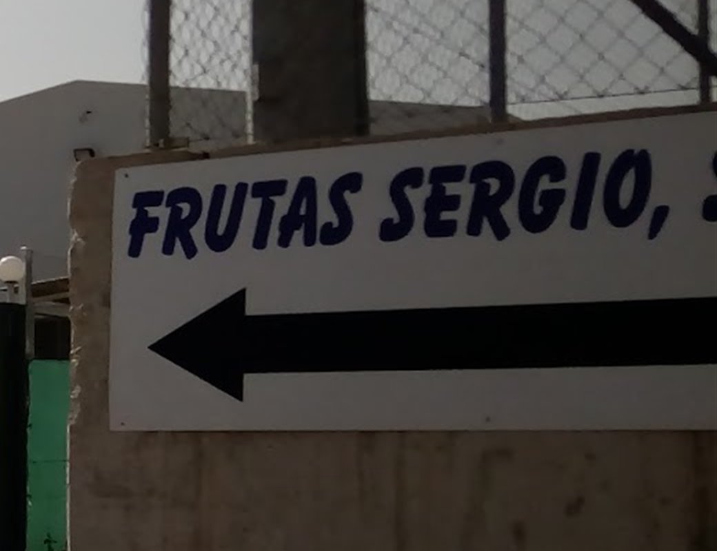 Cartel de Frutas Sergio con letras azules y una flecha negra sobre un muro de hormigón